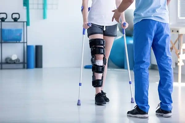 Patient in a knee brace walking with crutches, supported by a physiotherapist
